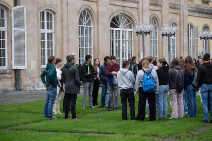 Foto: Eine Gruppe von Personen steht auf einem Rasen vor dem Schloss Hohenheim. Das Gebäude im Hintergrund hat große Fenster und klassische Architekturmerkmale. Die Personen tragen verschiedene Freizeitkleidung und scheinen in ein Gespräch vertieft zu sein.