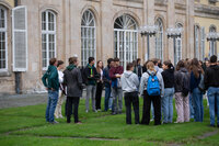 Foto: Eine Gruppe von Personen steht auf einem Rasen vor dem Schloss Hohenheim. Das Gebäude im Hintergrund hat große Fenster und klassische Architekturmerkmale. Die Personen tragen verschiedene Freizeitkleidung und scheinen in ein Gespräch vertieft zu sein.