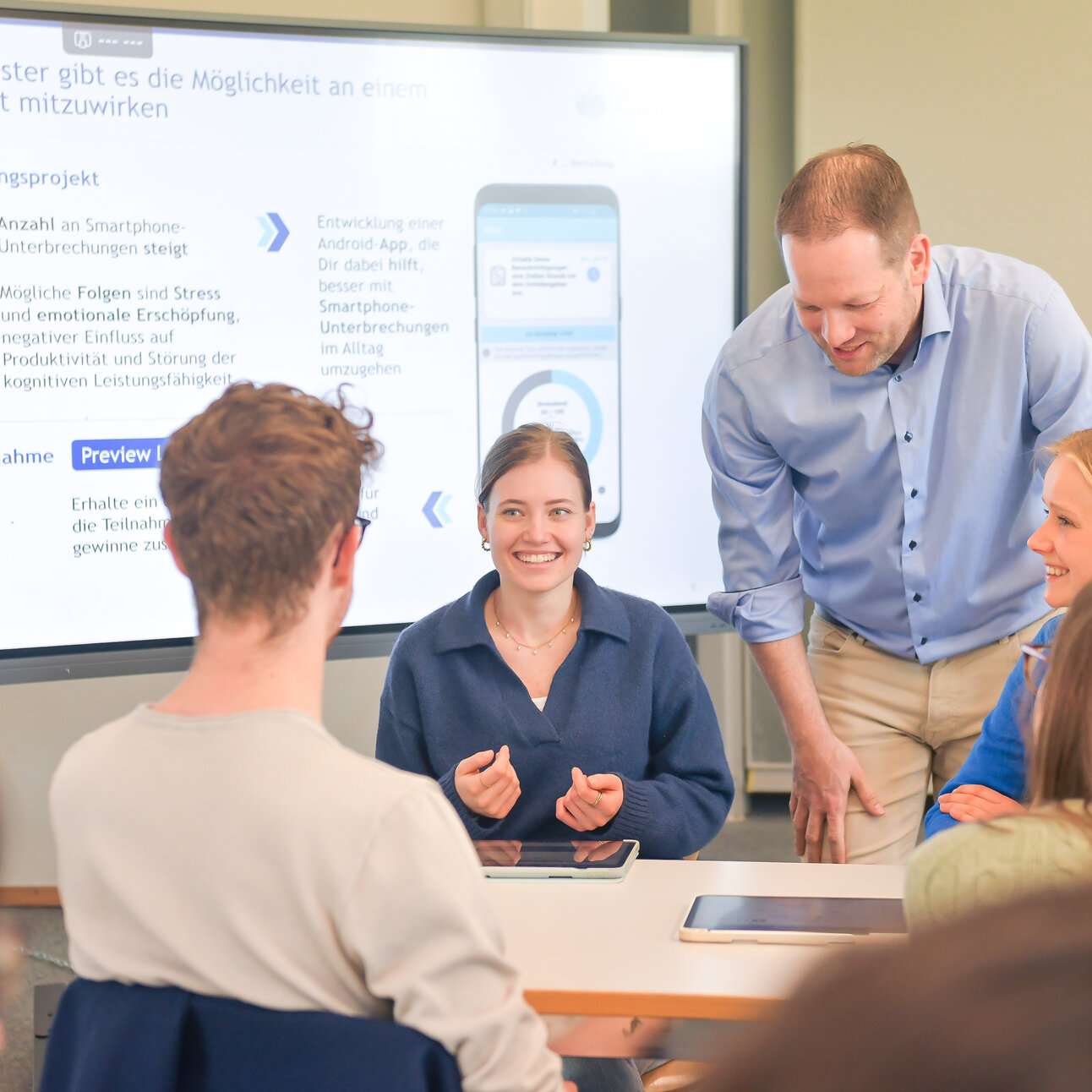 Foto: Eine Gruppe von Personen sitzt an einem Tisch und diskutiert ein Forschungsprojekt, das auf einem großen Bildschirm im Hintergrund angezeigt wird. Der Bildschirm zeigt Informationen über Smartphone-Unterbrechungen und deren Auswirkungen sowie eine App-Entwicklung. Eine Person steht und spricht mit den anderen.