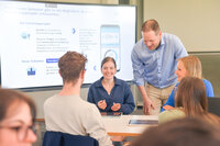 Foto: Eine Gruppe von Personen sitzt an einem Tisch und diskutiert ein Forschungsprojekt, das auf einem großen Bildschirm im Hintergrund angezeigt wird. Der Bildschirm zeigt Informationen über Smartphone-Unterbrechungen und deren Auswirkungen sowie eine App-Entwicklung. Eine Person steht und spricht mit den anderen.