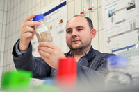 Foto: Eine Person in einem Labor hält eine Flasche mit einem blauen Deckel, die mit Probenmaterial gefüllt ist. Im Hintergrund sind Diagramme und Poster an der Wand sichtbar.