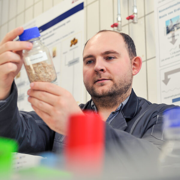 Foto: Eine Person in einem Labor hält eine Flasche mit einem blauen Deckel, die mit Probenmaterial gefüllt ist. Im Hintergrund sind Diagramme und Poster an der Wand sichtbar.