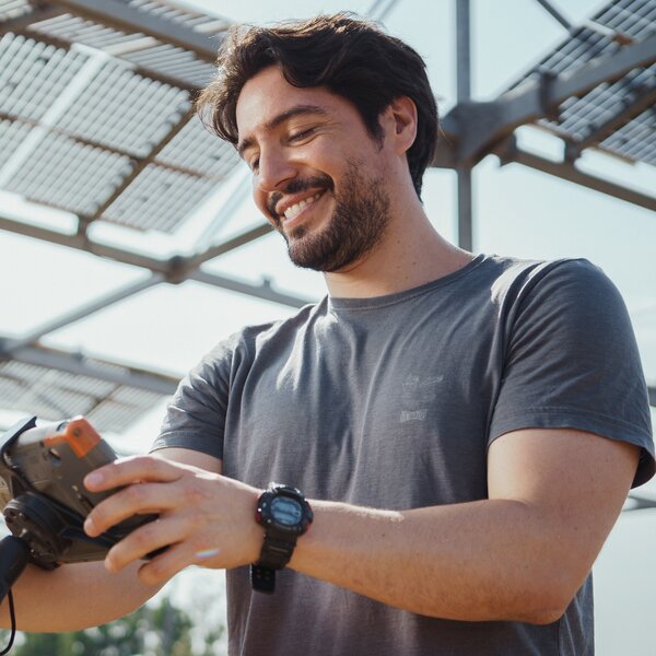 Foto: Eine Person hält ein Messgerät und steht unter einer Struktur mit Solarpanelen im Freien. Die Person trägt ein T-Shirt und eine Uhr.