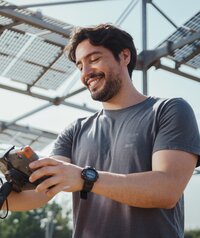 Foto: Eine Person hält ein Messgerät und steht unter einer Struktur mit Solarpanelen im Freien. Die Person trägt ein T-Shirt und eine Uhr.