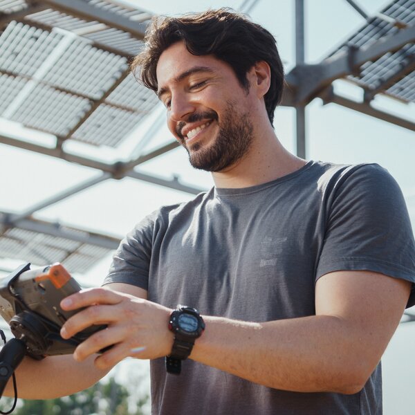Foto: Eine Person hält ein Messgerät und steht unter einer Struktur mit Solarpanelen im Freien. Die Person trägt ein T-Shirt und eine Uhr.
