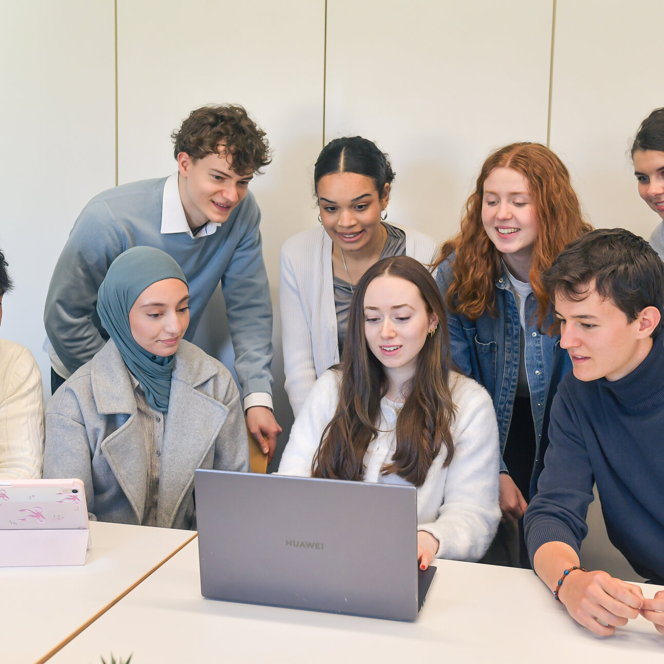 Foto: Eine Gruppe von Personen sitzt und steht um einen Tisch. Sie verwenden Laptops und Tablets. Die Personen sind in einem Raum mit weißen Wänden und tragen verschiedene Freizeitkleidung. Ein Laptop auf dem Tisch hat das Huawei-Logo.