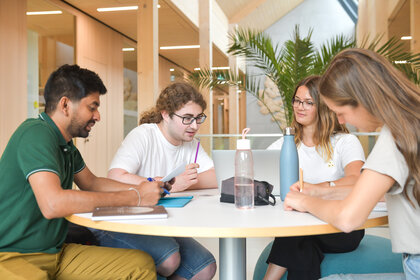 Foto: Vier Personen sitzen an einem runden Tisch in einem modernen Innenraum und arbeiten zusammen. Auf dem Tisch stehen Wasserflaschen und ein Laptop. Im Hintergrund sind Pflanzen und Holzstrukturen sichtbar.