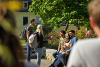 Foto: Eine Gruppe von Personen sitzt und steht im Freien vor einem Gebäude mit gelber Fassade. Zwei Personen mit Rucksäcken stehen, während drei Personen auf einer Bank sitzen und Getränke halten. Im Hintergrund sind blühende Sträucher und ein Baum zu sehen.