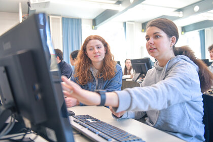 Foto: Mehrere Personen sitzen in einem Raum mit Computern. Eine Person zeigt auf den Bildschirm eines Computers, während eine andere Person zusieht. Im Hintergrund sind weitere Personen an Computern zu sehen.
