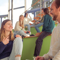 Foto: Mehrere Personen sitzen auf grünen Stufen in einem modernen, lichtdurchfluteten Raum mit großen Fenstern. Sie scheinen in ein Gespräch vertieft zu sein. Die Umgebung wirkt offen und einladend.