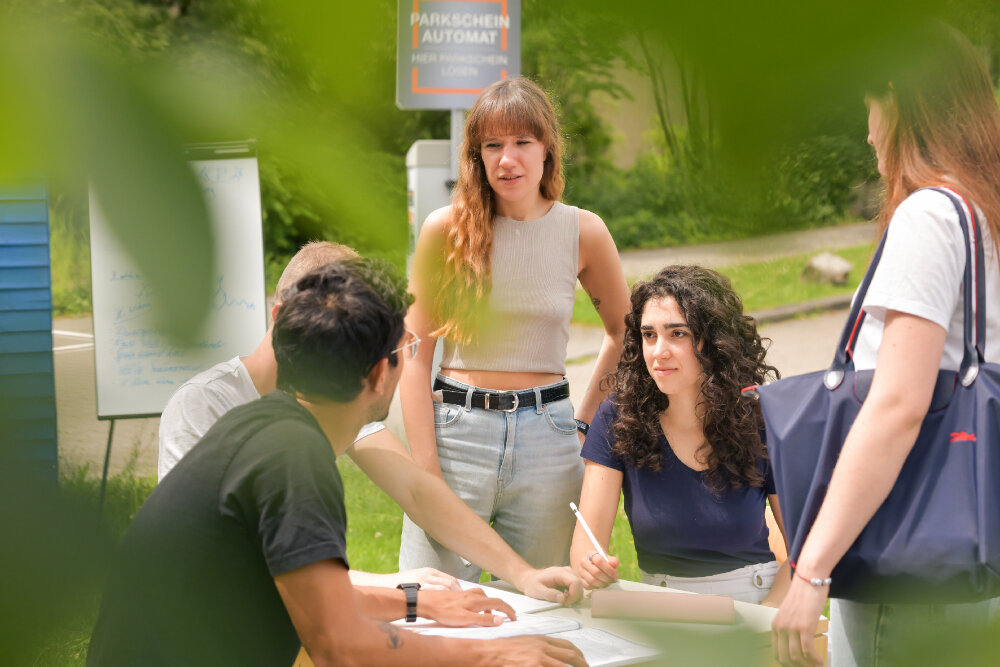 Foto: Eine Gruppe von Personen steht und sitzt um einen Tisch im Freien. Eine Person hält einen Stift und zeigt auf ein Dokument. Im Hintergrund ist ein Schild mit der Aufschrift "Parkschein Automat" sichtbar. Ein Whiteboard steht links. Die Szene ist von grünen Blättern umrahmt.