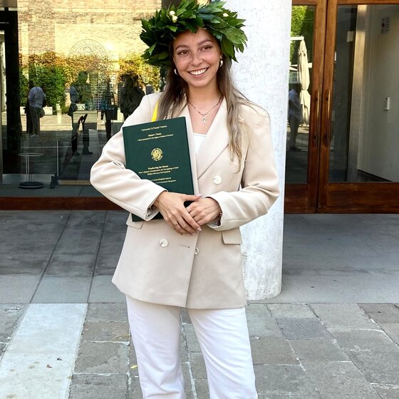 Eine Person steht vor einem Gebäude und trägt einen Lorbeerkranz. Sie hält ein Dokument mit dem Text "Università Ca' Foscari Venezia" und weiteren Details. Die Person trägt einen beigen Blazer und weiße Hose. Im Hintergrund ist ein Glasfenster und ein Eingang zu sehen.