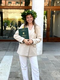 Eine Person steht vor einem Gebäude und trägt einen Lorbeerkranz. Sie hält ein Dokument mit dem Text "Università Ca' Foscari Venezia" und weiteren Details. Die Person trägt einen beigen Blazer und weiße Hose. Im Hintergrund ist ein Glasfenster und ein Eingang zu sehen.