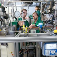Foto: Drei Personen in grünen Kitteln arbeiten in einem Labor an einer komplexen technischen Anlage mit Druckanzeigen und Schläuchen. Ein Display zeigt "7.02" und "38.2°C". Die Umgebung ist industriell mit zahlreichen Messgeräten und Kabeln.