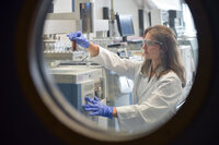 Foto: Eine Person in einem weißen Laborkittel arbeitet in einem Labor und bedient ein wissenschaftliches Gerät. Sie trägt blaue Handschuhe und hält ein Reagenzglas. Im Hintergrund sind weitere Laborgeräte und Computerbildschirme sichtbar.