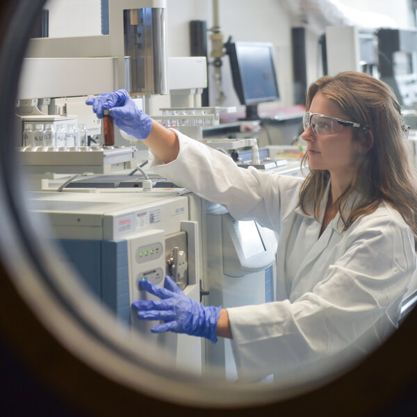 Foto: Eine Person in einem weißen Laborkittel arbeitet in einem Labor und bedient ein wissenschaftliches Gerät. Sie trägt blaue Handschuhe und hält ein Reagenzglas. Im Hintergrund sind weitere Laborgeräte und Computerbildschirme sichtbar.