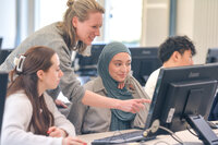 Lehrende zeigt drei Studierenden etwas am Bildschirm in einem Computerraum.