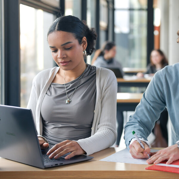 Foto: Zwei Personen sitzen an einem Tisch in einer Bibliothek. Eine Person arbeitet an einem Laptop, während die andere Notizen auf Papier macht. Im Hintergrund sind weitere Personen an Tischen sichtbar.