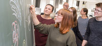 Foto: Eine Person schreibt mit Kreide an eine Tafel, während andere Personen zuschauen. Im Hintergrund ist ein Labor mit verschiedenen Geräten sichtbar.