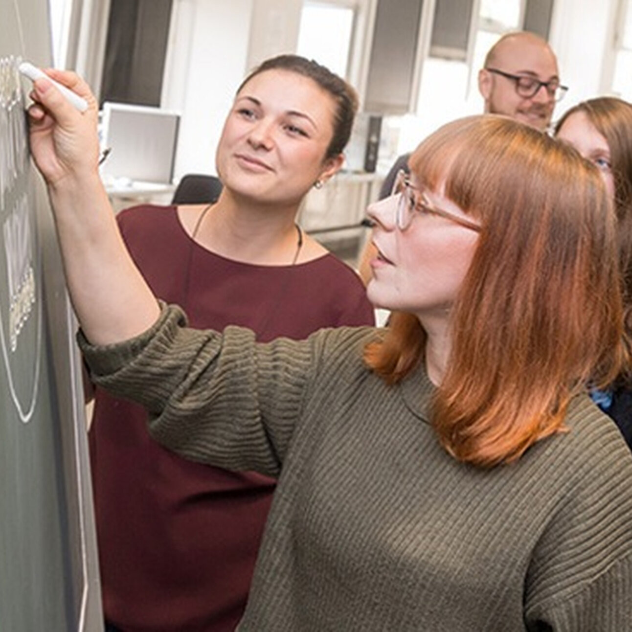Foto: Eine Person schreibt mit Kreide an eine Tafel, während andere Personen zuschauen. Im Hintergrund ist ein Labor mit verschiedenen Geräten sichtbar.