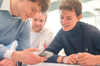 Foto: Drei Personen stehen zusammen und betrachten ein Smartphone. Eine Person hält das Gerät, während die anderen beiden zuschauen. Die Umgebung wirkt wie ein Innenraum.