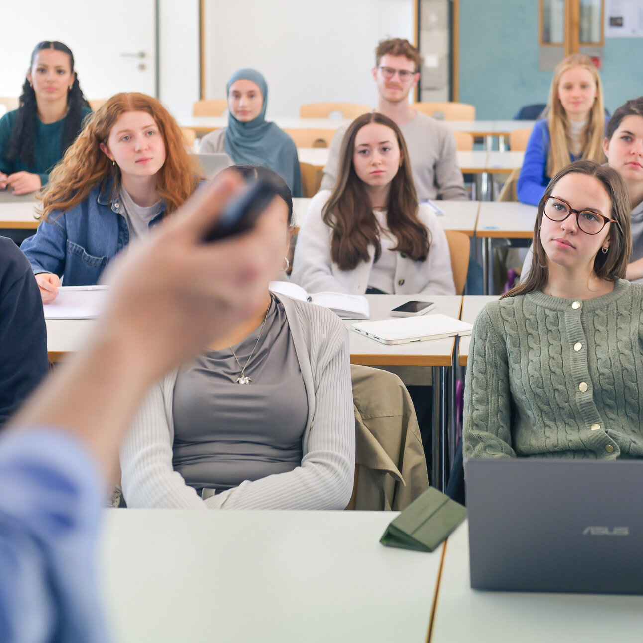Foto: Eine Gruppe von Personen sitzt in einem Hörsaal und hört einem Vortrag zu. Im Vordergrund ist eine Person mit einem Laptop zu sehen. Der Raum ist mit Tischen und Stühlen ausgestattet.