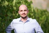 Eine Person in einem gestreiften Hemd steht vor einer Reihe von grünen Pflanzen. Die Umgebung wirkt natürlich und die Pflanzen scheinen Teil eines Gartens oder einer landwirtschaftlichen Fläche zu sein.