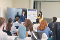 Zwei Studentinen präsentieren vor einer Gruppe mit Flipchart und großem Bildschirm.