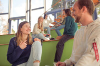 Foto: Mehrere Personen sitzen auf grünen Stufen in einem modernen, lichtdurchfluteten Raum mit großen Fenstern. Sie scheinen in ein Gespräch vertieft zu sein. Die Umgebung wirkt offen und einladend.