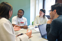 Foto: Vier Personen sitzen an einem Tisch und diskutieren. Eine Person trägt ein T-Shirt mit der Aufschrift 'BE BRAVE WITH YOUR LIFE'. Auf dem Tisch liegen Bücher und ein Laptop.
