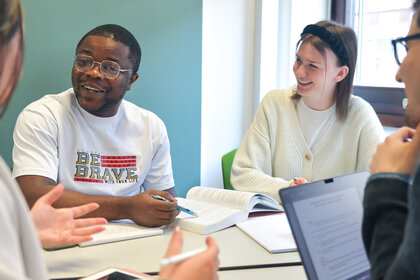 Foto: Vier Personen sitzen an einem Tisch und diskutieren. Eine Person trägt ein T-Shirt mit der Aufschrift 'BE BRAVE WITH YOUR LIFE'. Auf dem Tisch liegen Bücher und ein Laptop.