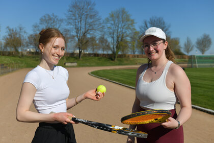 Foto: Zwei Personen stehen auf einem Tennisplatz im Freien. Eine Person hält einen Tennisball und einen Schläger, während die andere Person ebenfalls einen Schläger hält. Im Hintergrund sind ein gepflegter Rasen und Bäume sichtbar.