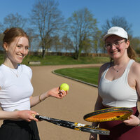 Foto: Zwei Personen stehen auf einem Tennisplatz im Freien. Eine Person hält einen Tennisball und einen Schläger, während die andere Person ebenfalls einen Schläger hält. Im Hintergrund sind ein gepflegter Rasen und Bäume sichtbar.