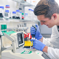 Foto: Eine Person in einem weißen Laborkittel arbeitet mit einer Pipette an einem Laborgerät. Im Hintergrund sind Regale mit Flaschen und Laborutensilien sichtbar.