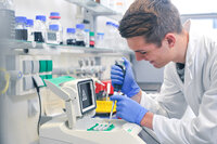 Foto: Eine Person in einem weißen Laborkittel arbeitet mit einer Pipette an einem Laborgerät. Im Hintergrund sind Regale mit Flaschen und Laborutensilien sichtbar.