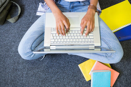 Foto: Eine Person sitzt auf einem Teppichboden mit überkreuzten Beinen und tippt auf einem Laptop. Neben der Person liegen bunte Notizbücher und eine Tasche. Die Person trägt Jeans und ein Armband.