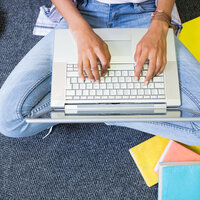 Foto: Eine Person sitzt auf einem Teppichboden mit überkreuzten Beinen und tippt auf einem Laptop. Neben der Person liegen bunte Notizbücher und eine Tasche. Die Person trägt Jeans und ein Armband.