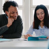 Foto: Zwei Personen sitzen an einem Tisch in einem hellen Raum. Eine Person trägt einen grauen Pullover, die andere eine hellblaue Bluse. Beide betrachten Dokumente auf dem Tisch.