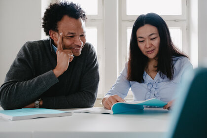 Foto: Zwei Personen sitzen an einem Tisch in einem hellen Raum. Eine Person trägt einen grauen Pullover, die andere eine hellblaue Bluse. Beide betrachten Dokumente auf dem Tisch.