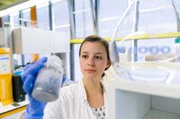 Foto: Eine Person in einem Labor hält eine Flasche mit unleserlichem Text. Die Person trägt einen weißen Laborkittel und blaue Handschuhe. Im Hintergrund sind Laborgeräte und Fenster zu sehen.