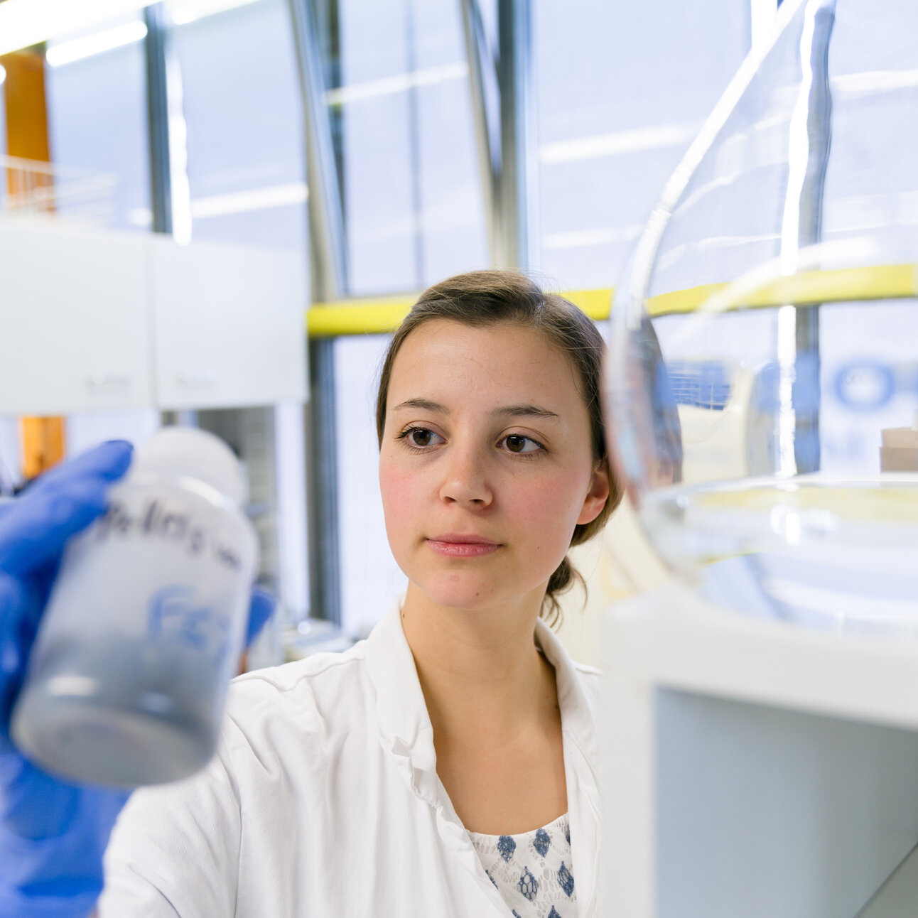 Foto: Eine Person in einem Labor hält eine Flasche mit unleserlichem Text. Die Person trägt einen weißen Laborkittel und blaue Handschuhe. Im Hintergrund sind Laborgeräte und Fenster zu sehen.