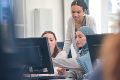 Foto: Drei Personen arbeiten zusammen an Computern in einem Büro. Eine Person zeigt auf den Bildschirm, während die anderen aufmerksam schauen. Monitore und Büroeinrichtung sind sichtbar.