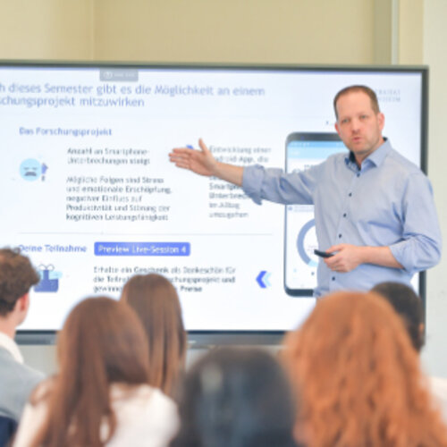 Eine Person steht vor einer Gruppe und präsentiert Informationen auf einem großen Bildschirm. Der Text auf dem Bildschirm beschreibt ein Forschungsprojekt mit Details zur Teilnahme. Im Hintergrund sind mehrere sitzende Personen zu sehen, die der Präsentation folgen.