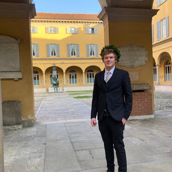 Eine Person in einem Anzug steht unter einem Bogengang vor einem historischen Gebäude mit gelber Fassade und blauen Fensterläden. Im Hintergrund ist ein Innenhof mit einer Statue sichtbar. Tafeln mit Inschriften sind an den Wänden angebracht.