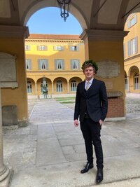 Eine Person in einem Anzug steht unter einem Bogengang vor einem historischen Gebäude mit gelber Fassade und blauen Fensterläden. Im Hintergrund ist ein Innenhof mit einer Statue sichtbar. Tafeln mit Inschriften sind an den Wänden angebracht.