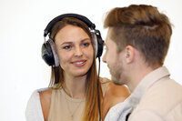 Foto: Zwei Personen stehen sich gegenüber. Eine Person trägt Kopfhörer und ein ärmelloses Oberteil. Die andere Person ist teilweise sichtbar. Der Hintergrund ist neutral und hell.