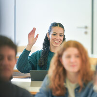 Foto: Mehrere Personen sitzen in einem Klassenzimmer. Eine Person hebt die Hand, während andere aufmerksam sind. Im Hintergrund sind Tische und Stühle sichtbar.