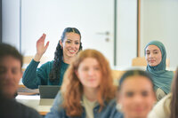 Foto: Mehrere Personen sitzen in einem Klassenzimmer. Eine Person hebt die Hand, während andere aufmerksam sind. Im Hintergrund sind Tische und Stühle sichtbar.