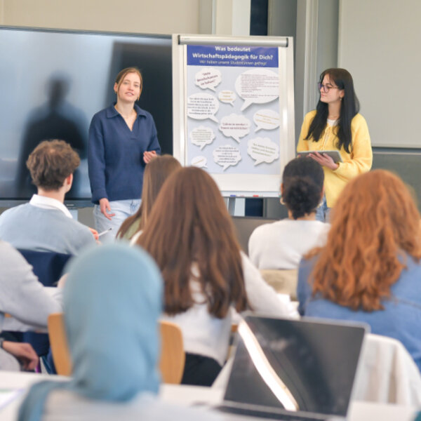 Zwei Studentinen präsentieren vor einer Gruppe mit Flipchart und großem Bildschirm.