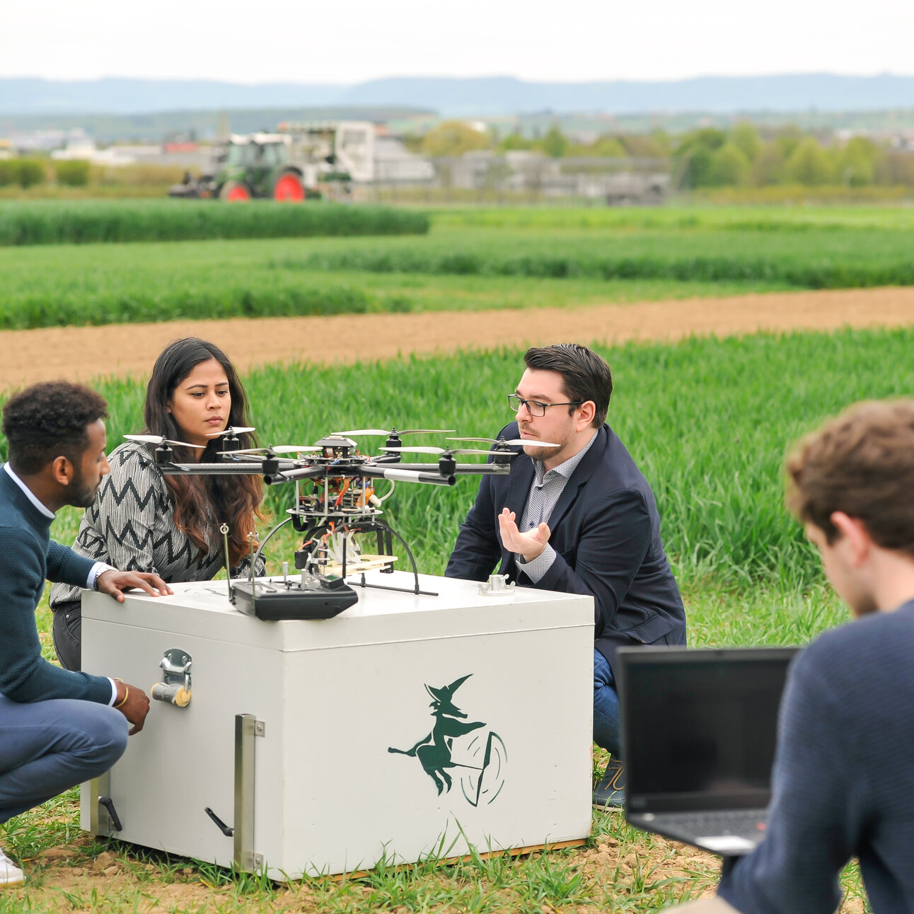 Foto: Vier Personen sind auf einem Feld zu sehen, die um eine große Drohne herum stehen. Eine Person hält einen Laptop, während die anderen die Drohne betrachten und diskutieren. Im Hintergrund sind Felder und ein Traktor sichtbar.